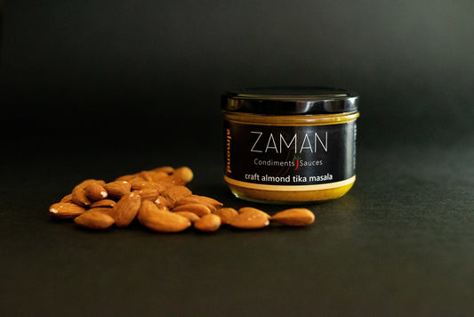 A glass jar with whole almonds next to it. The jar reads Zaman Craft Almond Tika Masala. The background is black and the word almond in a beige colour is written vertically on the label.
