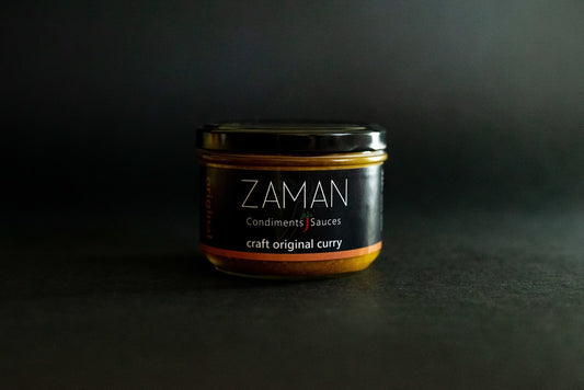 A glass jar with a black label. The label reads Zaman Condiments and Sauces and has a little red chilli in between the words condiments and sauces. The background is dark. The label reads craft original curry and there is the word original written vertically in brown.