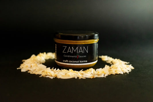 A glass jar with organic coconut next to it. The jar reads Zaman Craft Coconut Korma. The background is black and the word coconut in a white colour is written vertically on the label. The background is dark and the logo on the label reads Zaman condiments and sauces.