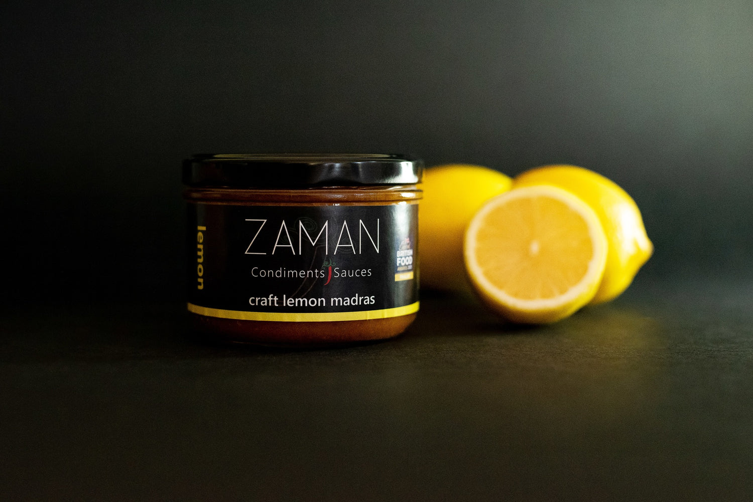 A glass jar with a black label. The label reads Zaman Condiments and Sauces and has a little red chilli in between the words condiments and sauces. The background is dark and there are 3 yellow lemons next to the jar.. The label reads craft lemon madras and there is the word lemon written vertically in yellow on the left of the label.